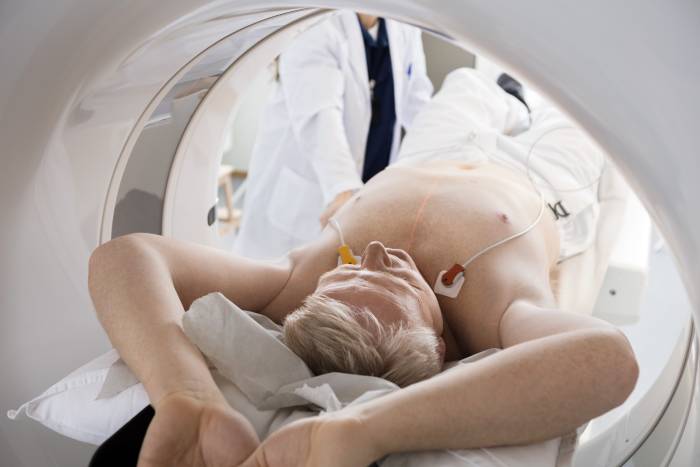 Close up view of a CT machine with an elderly man lying with outstretched hands and a radiologist taking him into the CT machine.